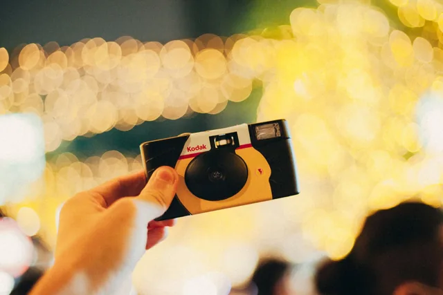 Une main tient un appareil photo jetable devant un fond de guirlandes lumineuses floues